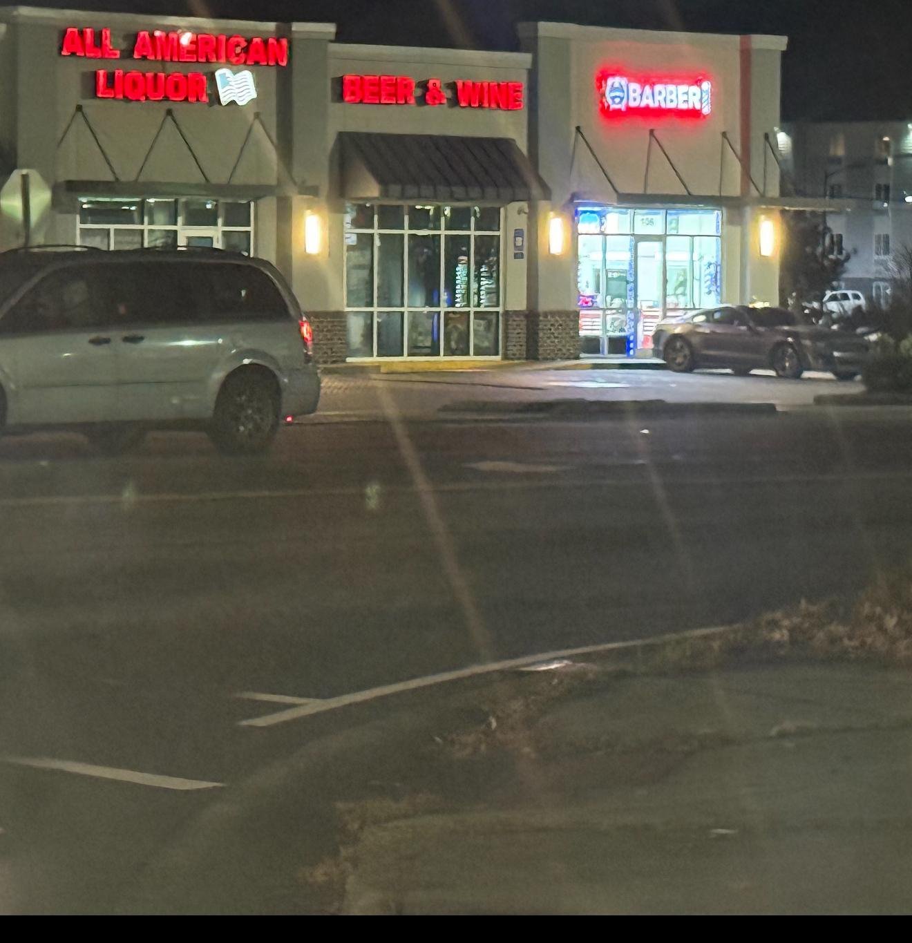 Night view of strip mall storefronts with red neon signs including All American Heroes, Beef and Wine, and Barger, with parked cars visible in foreground